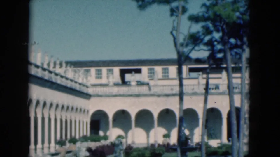 Wide 1975 Florida home-movie frame showing an arched arcade, upper facade, tall trees, garden shrubs, and classical courtyard details.
