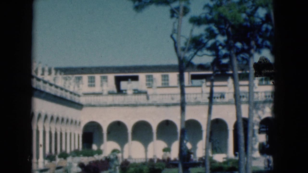 1975 Florida 8mm frame showing a sunlit stucco building with arches, balcony railings, pine trunks, and landscaped grounds.