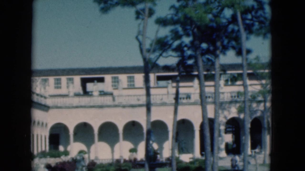 Arched building facade with balcony, pine trunks, garden area, and distant people.