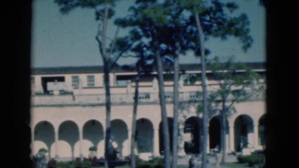 Soft 8mm view of a Florida building exterior in 1975, with pine trunks crossing a balcony and arched ground-floor walkway.