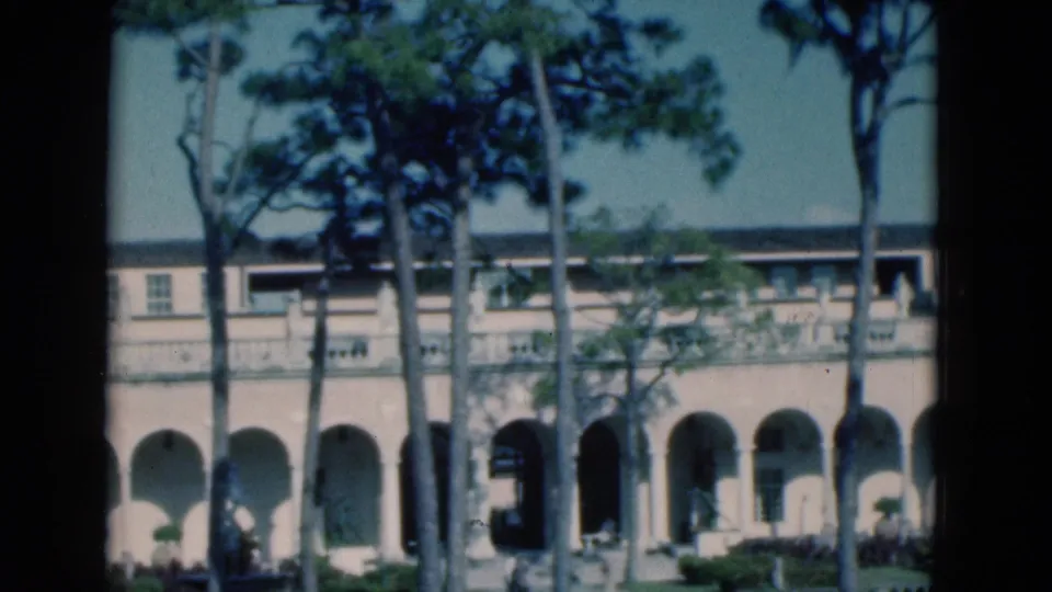 1975 Florida 8mm frame of an arched stucco exterior and balcony behind trees, with landscaped grounds and tiny distant people visible.