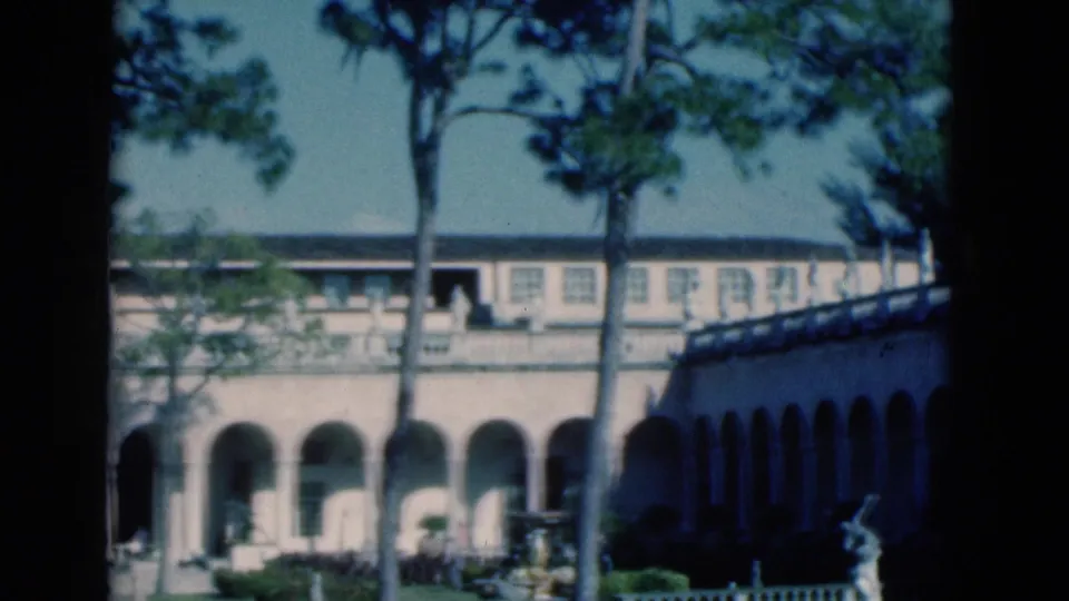 1975 Florida frame with arched building facade, tall pines, landscaped garden, and a small sculpture visible near the foreground.