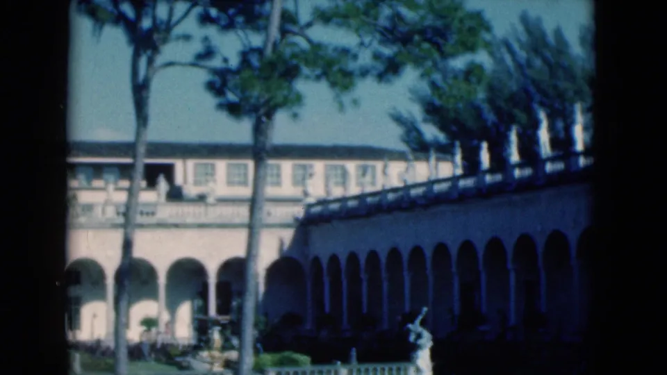 1975 Florida 8mm frame showing the colonnaded building, a row of side arches, palms or pines, and a garden sculpture.