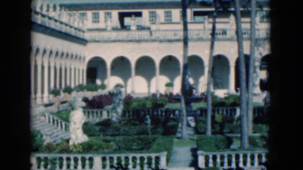 Formal mansion courtyard with arched colonnade, palms, hedges, statue, and white balustrade.