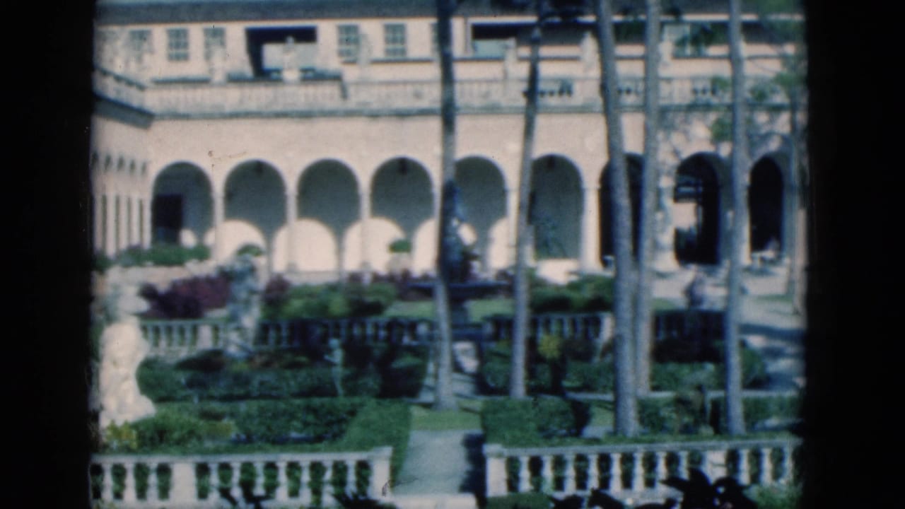 Mansion courtyard with arched facade, garden paths, palms, hedges, and distant people.