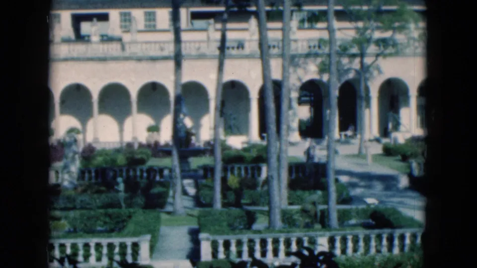 1975 Florida 8mm frame with a closer view of mansion arches, palm trunks, garden walkway, clipped planting, and white railing.