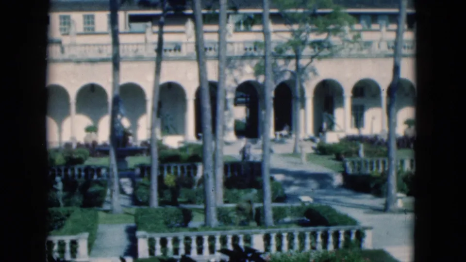 1975 Florida home-movie frame of a mansion courtyard path, arched colonnade, palms, hedges, balcony rail, and balustrade.