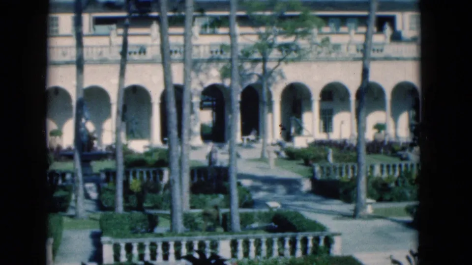 1975 Florida 8mm frame showing a landscaped courtyard with arched colonnades, palm trunks, garden paths, hedges, and a balustrade.