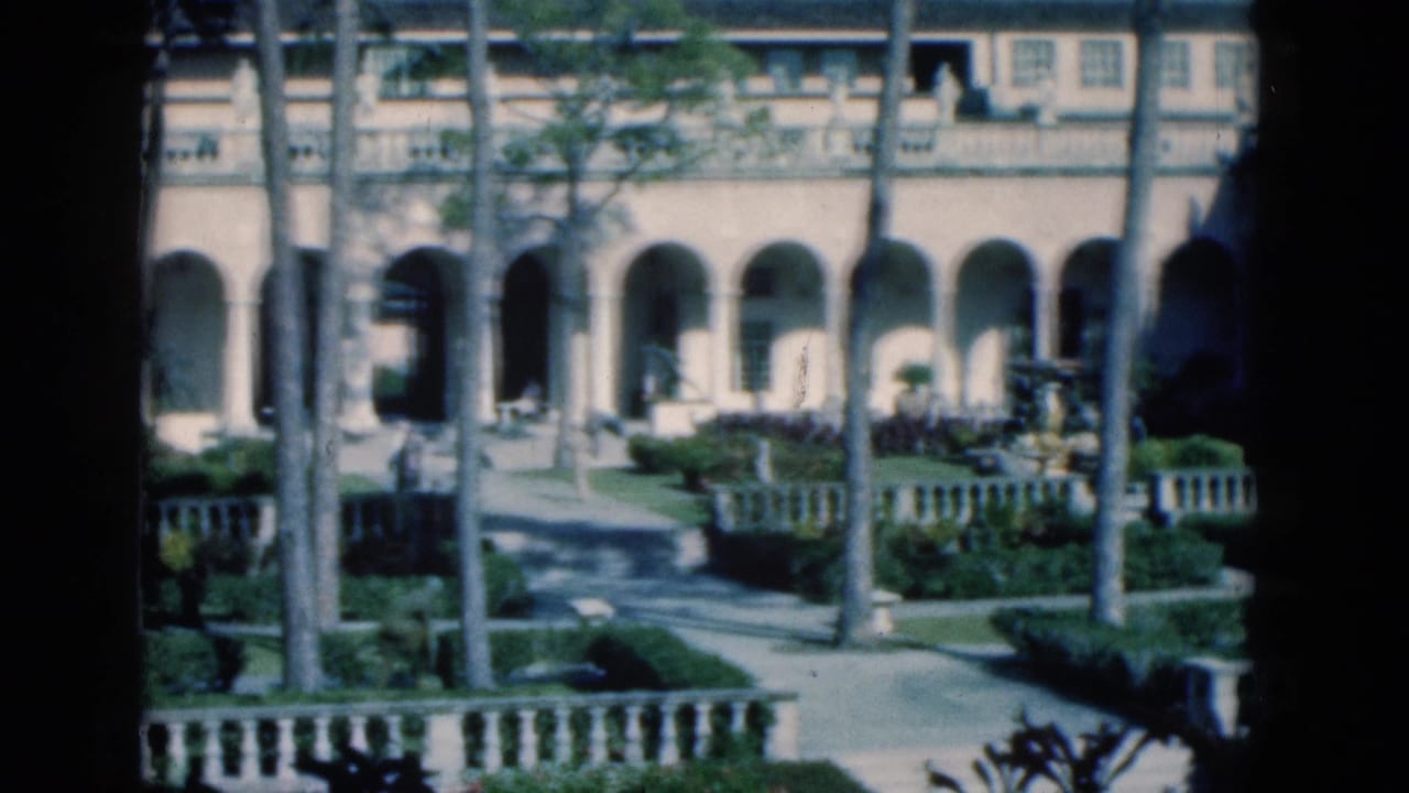 Arched building facade behind palms, hedges, pathways, and low stone garden railings.