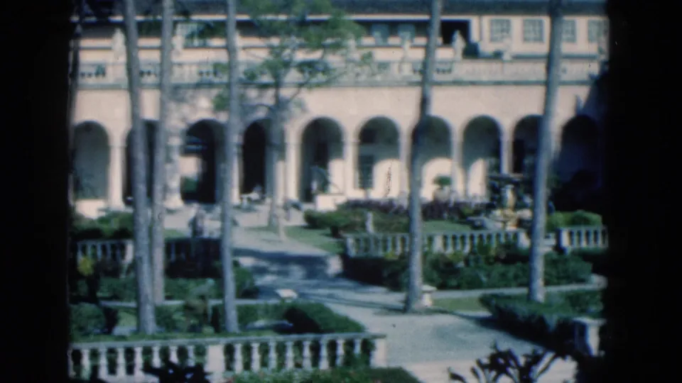 1975 Florida 8mm view of an arched courtyard facade with palms, clipped landscaping, paved paths, and low stone railings.
