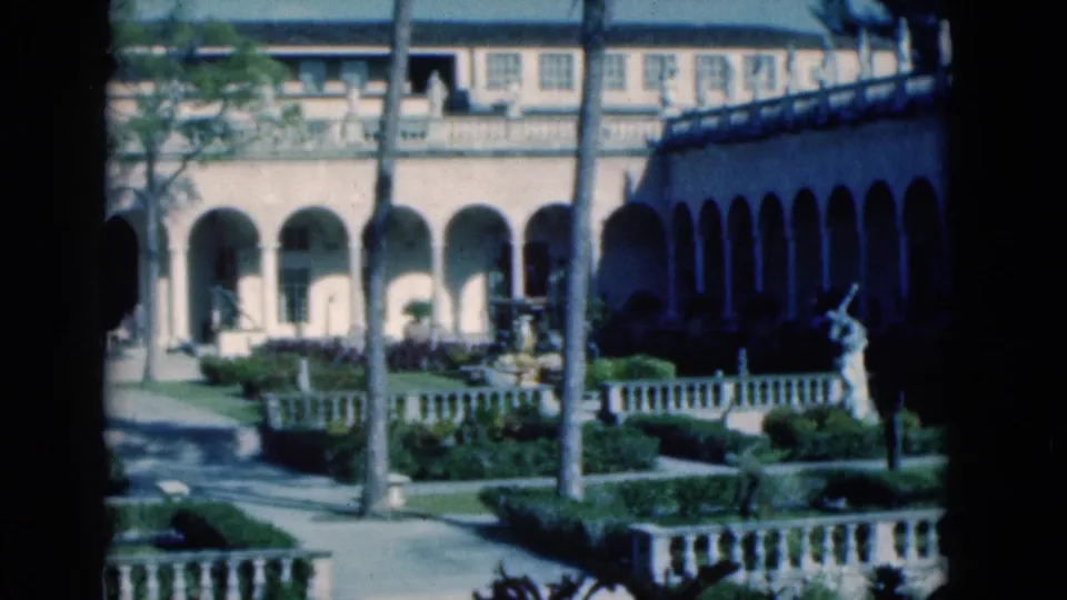 1975 Florida 8mm frame with a landscaped courtyard, arched colonnades, palm trunks, stone railings, and a small statue or fountain form.