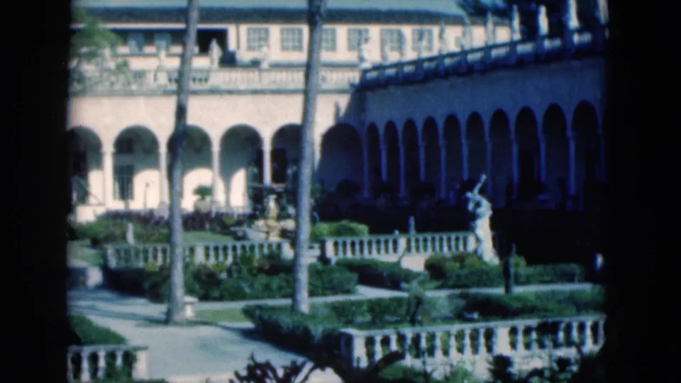 1975 Florida 8mm view of a palm-lined courtyard with arched walkway, clipped hedges, stone balustrade, and a small statue or fountain.