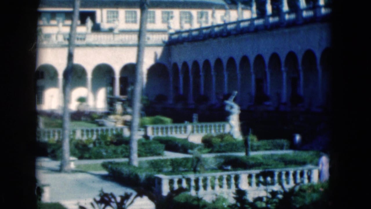 1975 Florida 8mm frame showing a sunlit arched courtyard with palms, manicured planting, stone railings, and a classical statue.