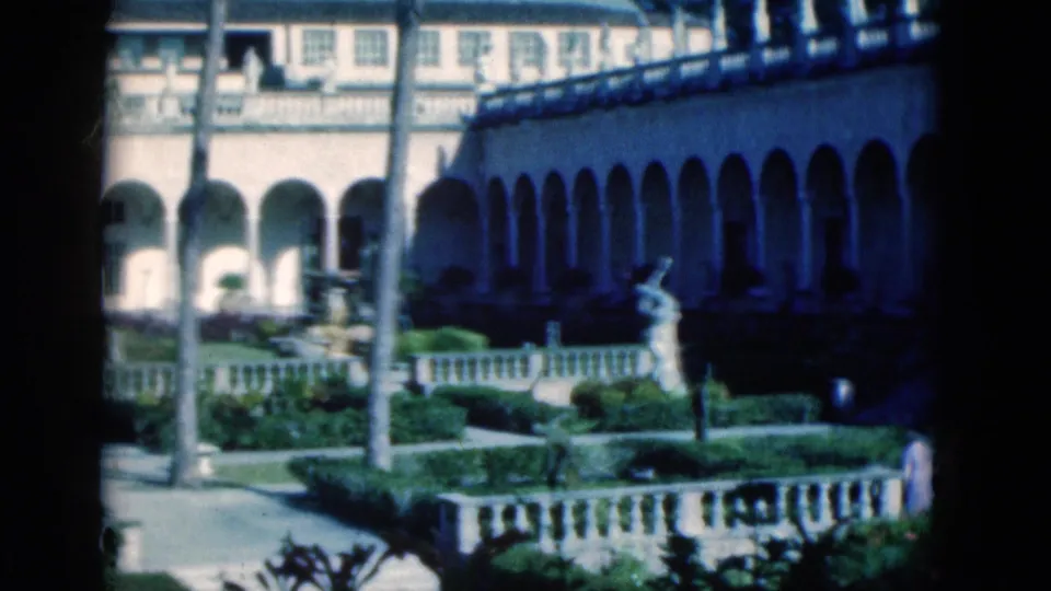 1975 Florida 8mm frame showing a sunlit arched courtyard with palms, manicured planting, stone railings, and a classical statue.