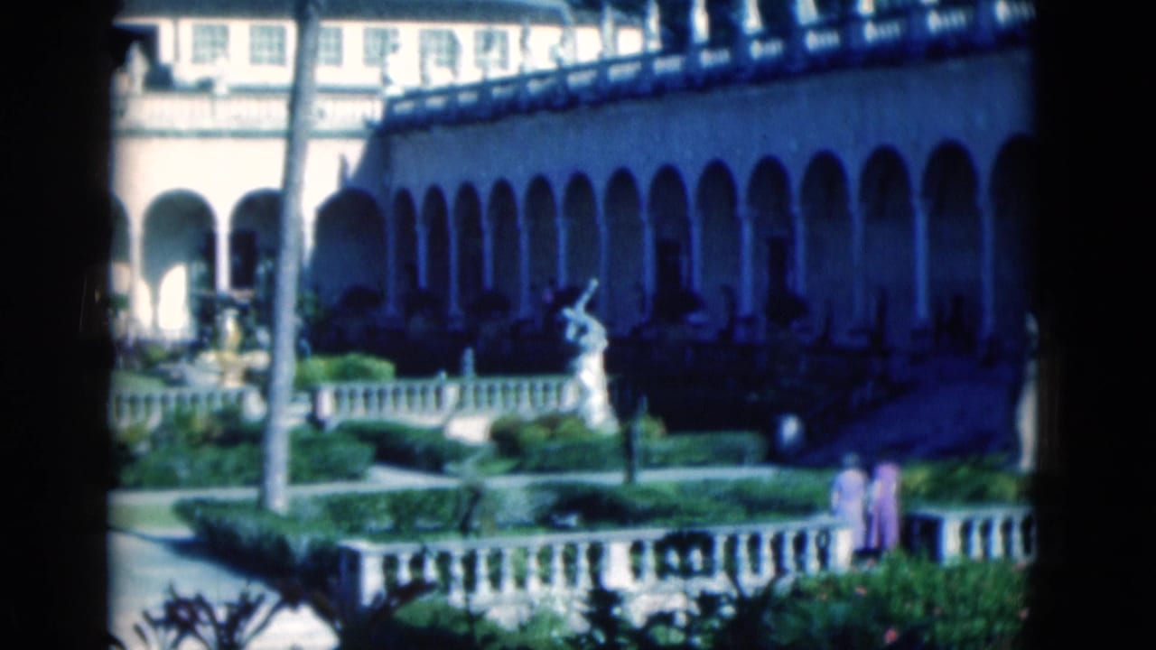 Courtyard garden with arched building, statue, palms, and two adult visitors.