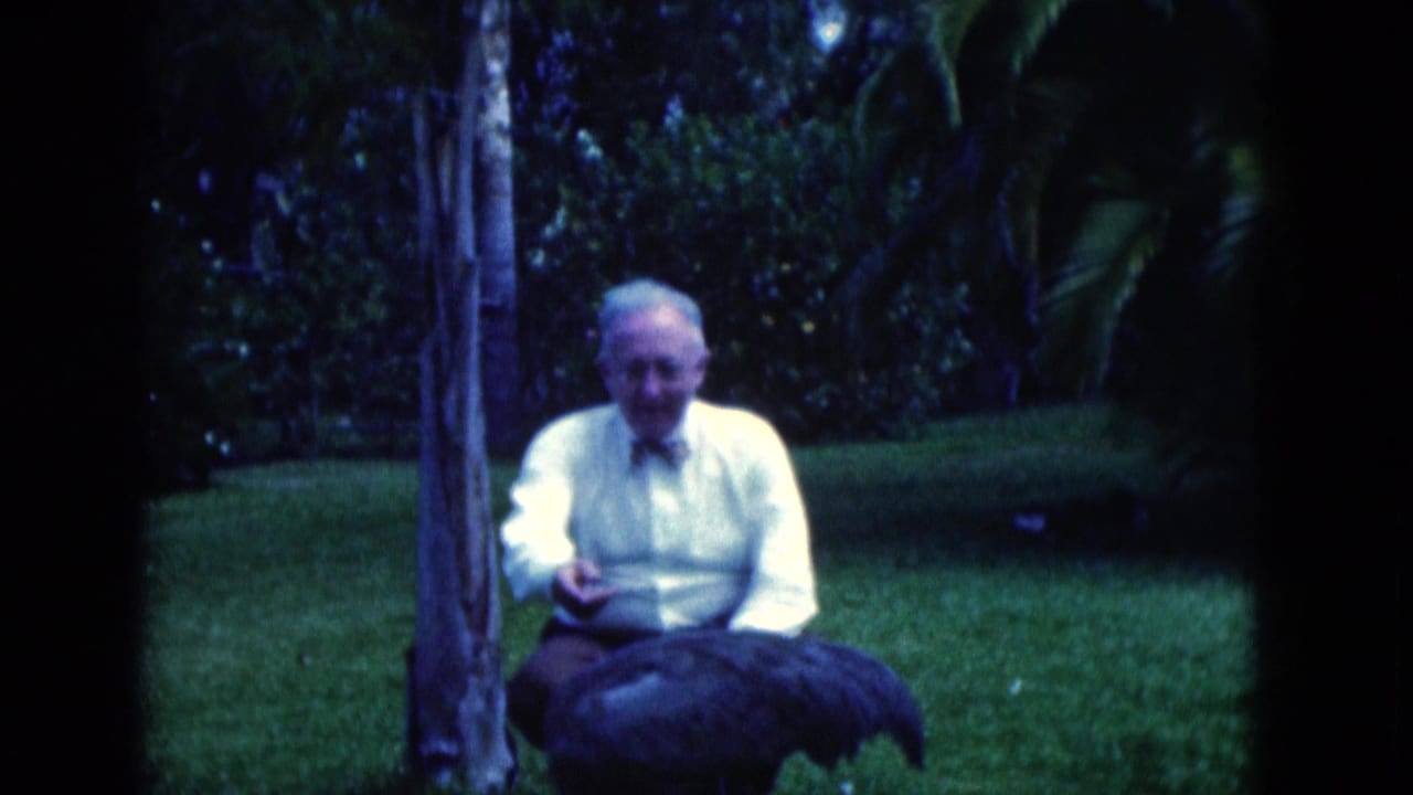 Older man seated on grass beside a large dark bird in a garden.
