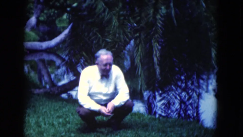 In soft 8mm color, an older man crouches on a Florida garden lawn beside palm trunks and shadowed foliage.