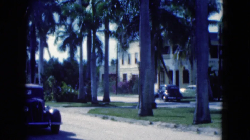 1975 Florida 8mm frame showing a curving palm-lined street, a dark vintage car, lawns, and pale low-rise buildings.