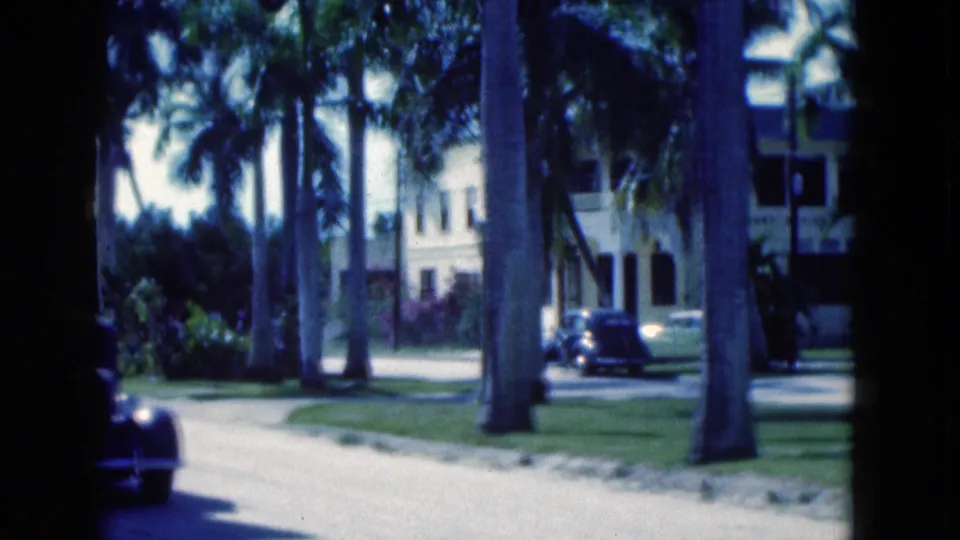 1975 Florida home-movie frame of a dark car on a palm-lined road beside lawns and pale low-rise buildings.