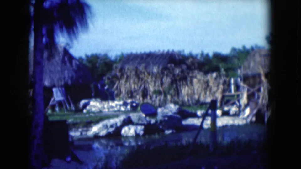 1975 Florida home-movie frame of thatched huts and a straw-covered wall behind rocks, water, palms, and greenery.