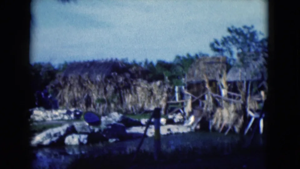 1975 Florida 8mm frame showing an empty rustic hut area with rough wooden supports, pale rocks, dark water, and dense foliage.