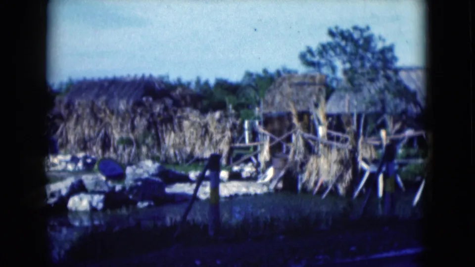1975 Florida frame of overgrown thatched structures, timber railings, palms, rocks, and a dark foreground water feature, with no people visible.