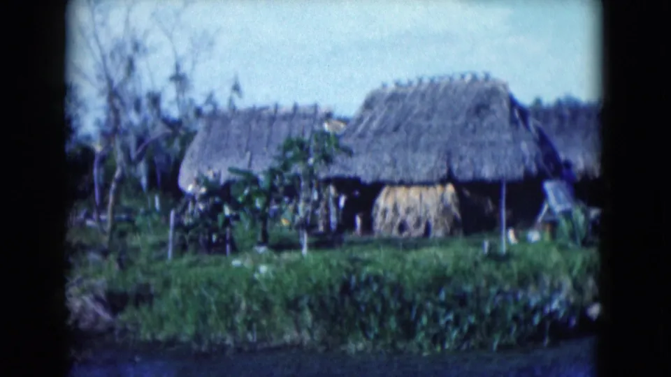 1975 Florida 8mm frame showing thatched huts, a straw stack, subtropical plants, and a calm foreground waterway.