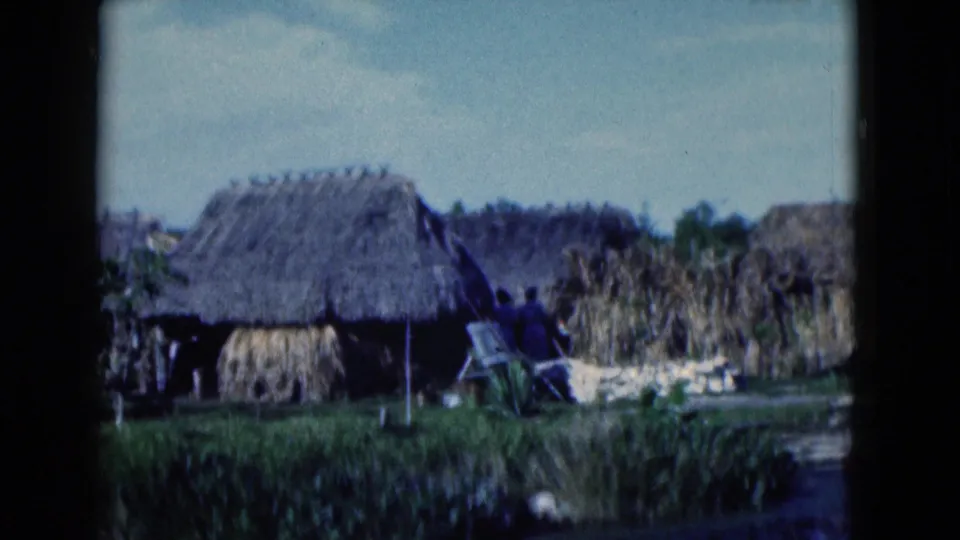 1975 Florida home-movie frame of thatched huts, reed stacks, marsh grass, and a foreground waterway under bright daylight.