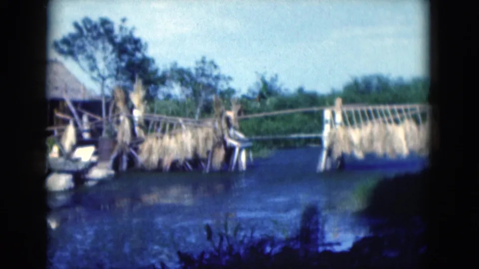 1975 Florida 8mm frame of a rough wooden bridge over water with dried plant material hanging from the structure.