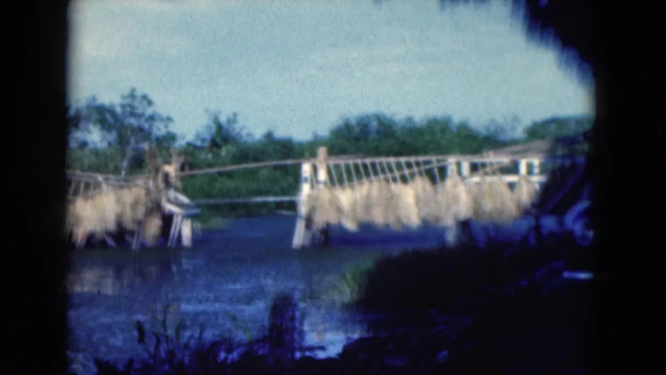 1975 Florida 8mm frame of a waterway beside a handmade bridge with dried plant bundles and surrounding greenery.