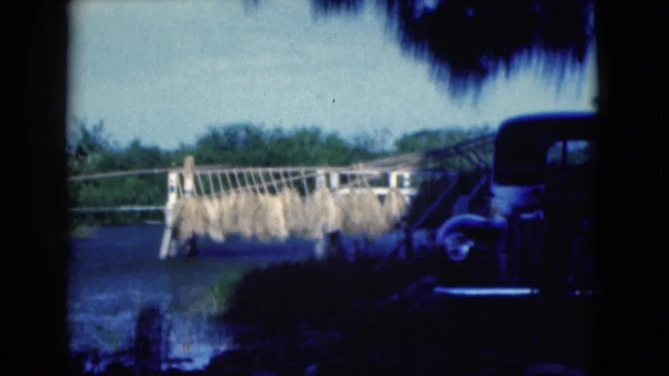 1975 Florida home-movie frame of drying crops hanging from a wooden structure, with a dark classic car partly framed at right.