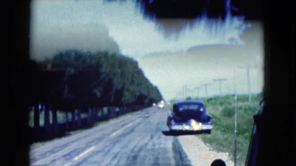 1975 Florida 8mm roadside view with a dark classic car on the shoulder, trees lining the road, and utility poles receding into the distance.
