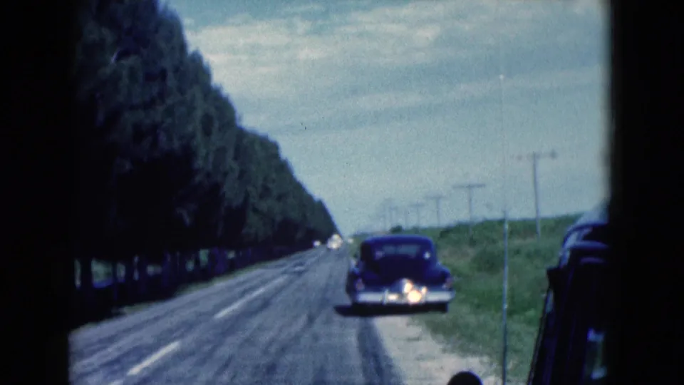 1975 Florida home-movie frame showing a dark classic car on a quiet rural road, bordered by trees, grass, utility poles, and open sky.