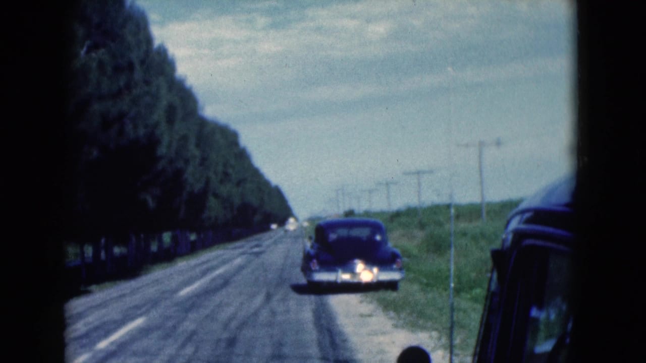 1975 Florida 8mm frame looking down a two-lane road past utility poles and a dark sedan stopped on the shoulder.