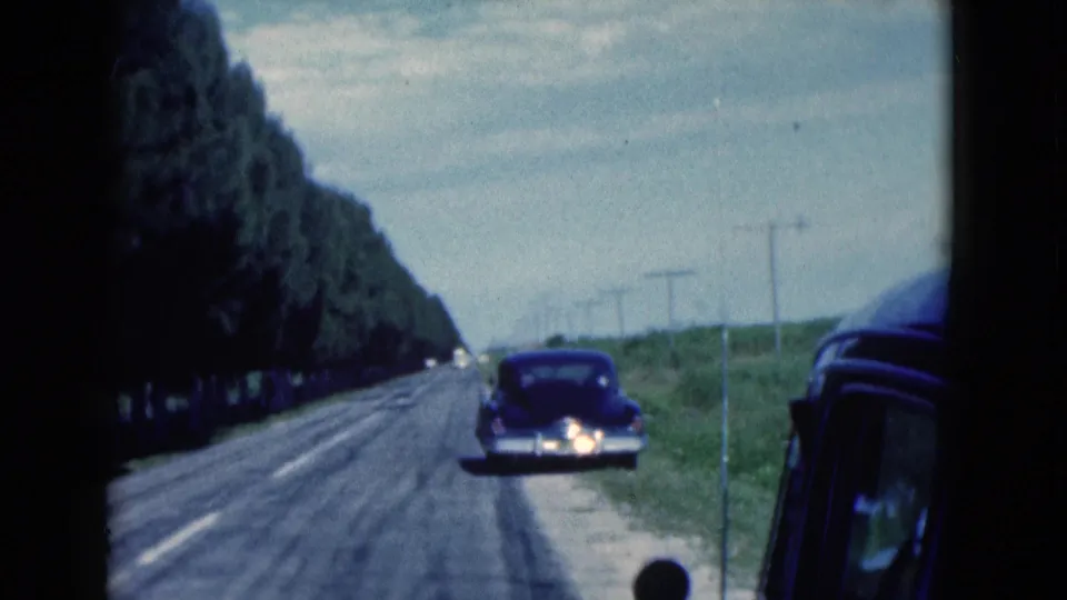 A rural Florida roadside view shows a dark classic sedan stopped on the shoulder along a two-lane road in 1975 home-movie footage.