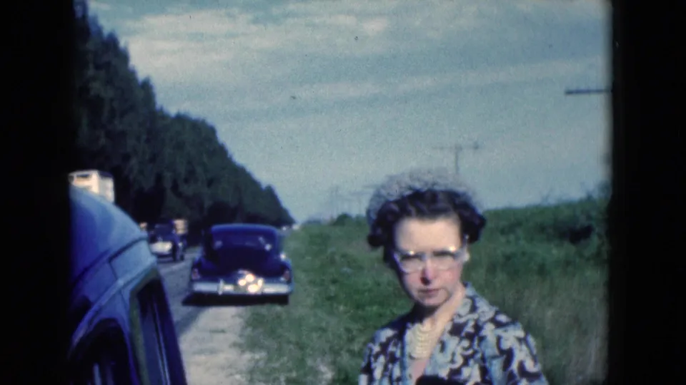 A woman wearing glasses and a patterned dress stands beside the Florida roadside as classic cars appear behind her.
