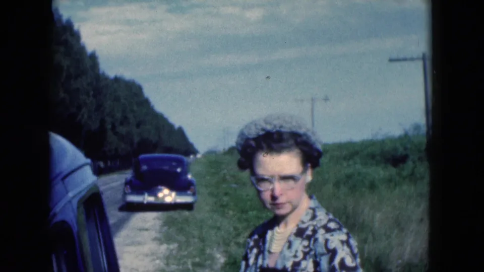 The final 1975 Florida frame shows a woman in glasses beside the grassy shoulder, with a dark classic sedan and utility poles behind her.
