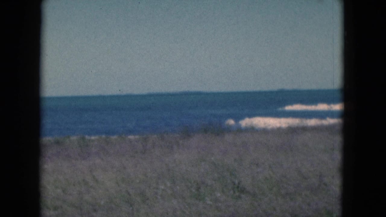 Grassy dune foreground with ocean and pale surf beyond, framed by dark film edges.