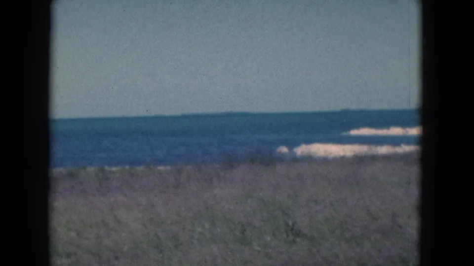 1975 Florida 8mm frame showing dune grass, blue ocean, pale surf, and a distant sandy shoreline under clear sky.