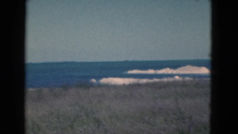 1975 Florida 8mm shoreline view with muted colors, dune grass below, ocean beyond, and pale waves breaking near sand.