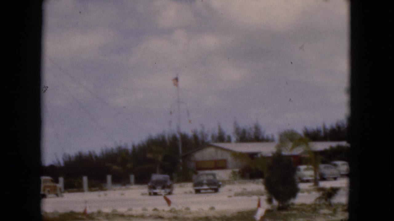 1975 Florida 8mm frame showing a low coastal building, several parked cars, a flagpole, trees, and cloudy sky.