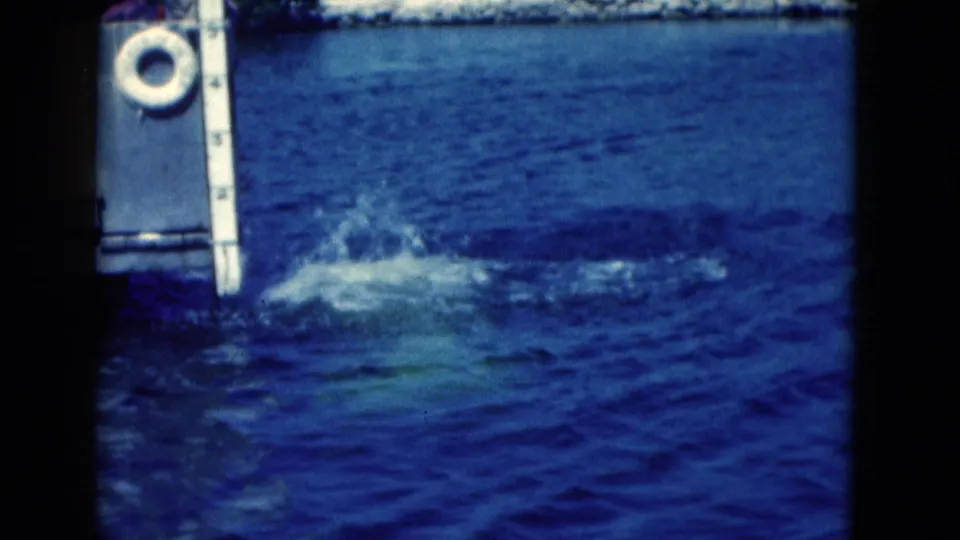 1975 Florida 8mm frame showing blue water splashing beside dock hardware, with a white ring fixture and vertical measuring post visible.