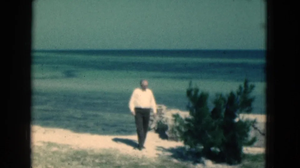 1975 Florida 8mm frame of an adult walking along a sandy beach near low shrubs, with blue-green coastal water behind him.