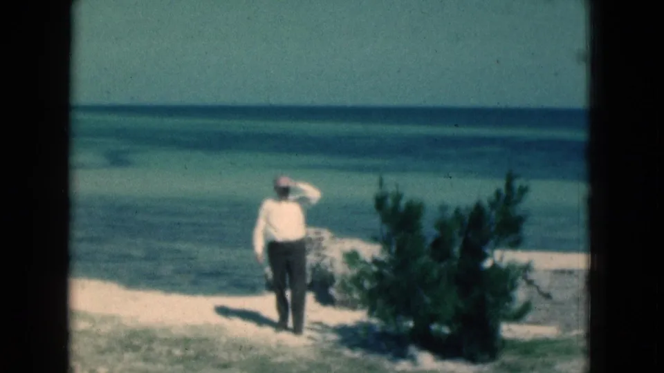The 1975 Florida beach scene shows the man standing near a low shrub, hand at his forehead, with layered ocean tones beyond.