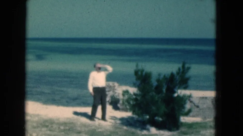 At the end of the 1975 Florida clip, the man remains by the shrub with his hand raised, facing the sunlit ocean.