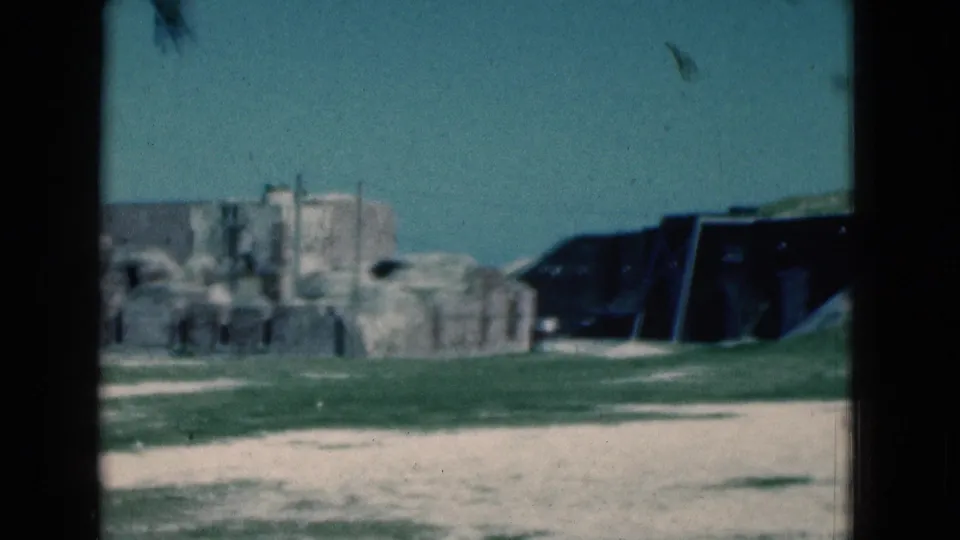 Soft 1975 Florida 8mm frame of weathered stone walls, open grass, a pale path, and a dark angular structure in a coastal historic-site view.