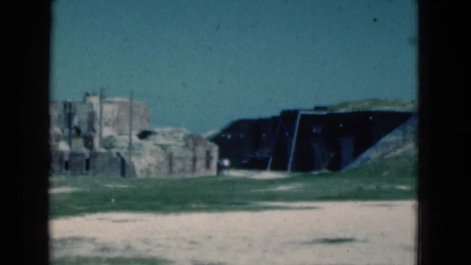 1975 Florida 8mm frame with partial stone ruins, a dark bunker-like building, grass, and a sandy path in a coastal sightseeing scene.