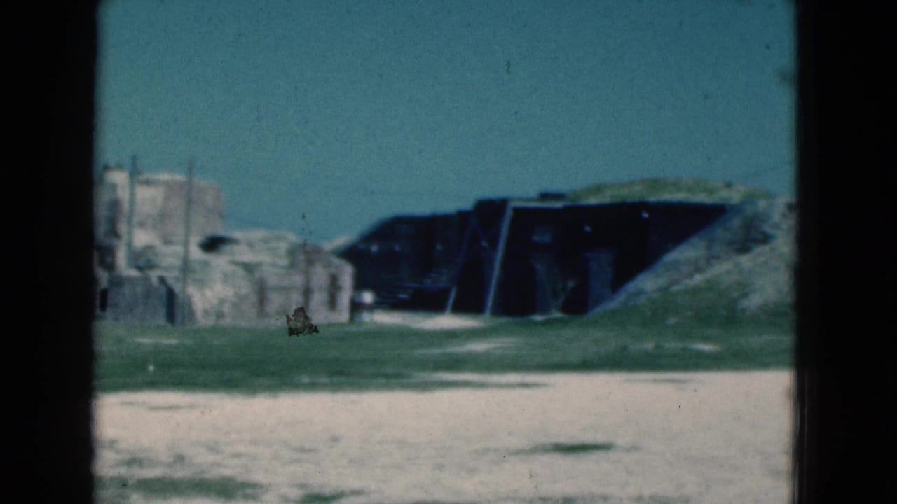 1975 Florida 8mm frame showing sandy foreground, grass, a pale small structure, and dark low openings set into mounds.