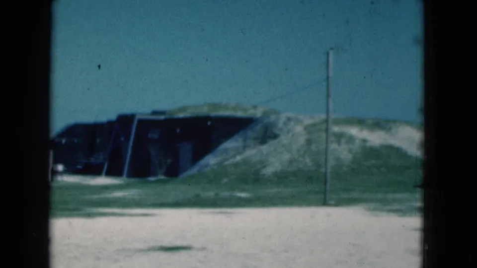 1975 Florida 8mm frame showing a dark low opening in a sandy mound, green field, pale ground, and a telephone pole.