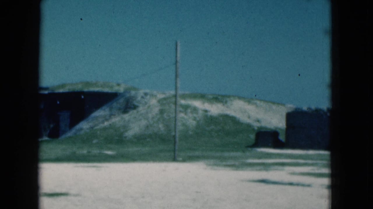 1975 Florida 8mm frame showing a grassy shoreline area with a low mound, a dark structure-like form, and open sky.